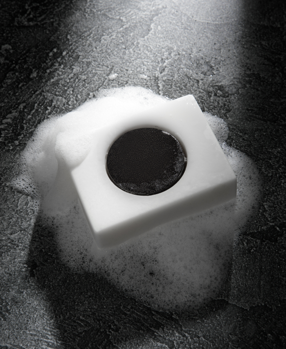 White soap bar with a circular black center surrounded by soap suds on a dark textured surface
