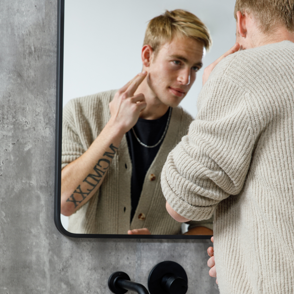 Young man with tattoo wearing beige cardigan examining his skin in bathroom mirror, skincare routine concept