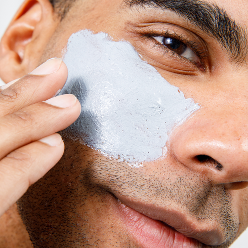 Man applying gray clay facial mask on cheek for skincare, grooming, and men's beauty routine close-up
