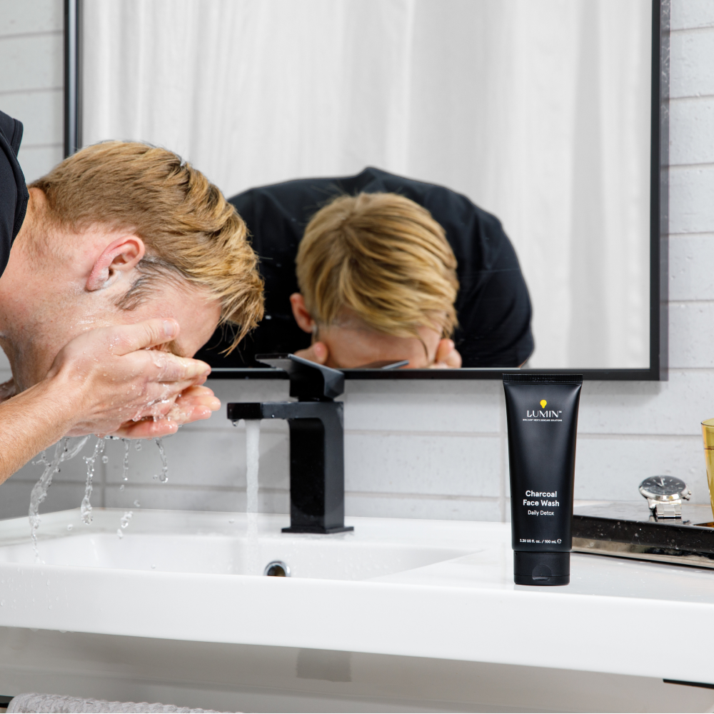Man washing face with water next to Lumin charcoal face wash tube on modern white bathroom sink with black faucet.
