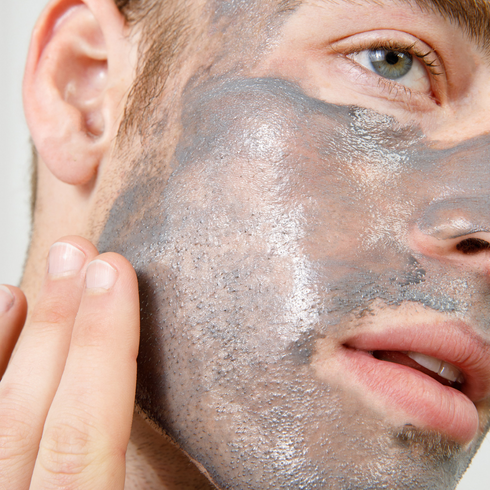 Man applying gray clay face mask on cheek with fingers, skincare routine, facial treatment for men, close-up.