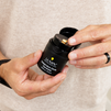 Hands holding Lumin Instant Dark Circle Corrector black container with beige concealer cream on finger, men's skincare product.