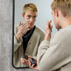 Young man applying green face mask cream from black skincare jar in front of mirror, men's grooming routine