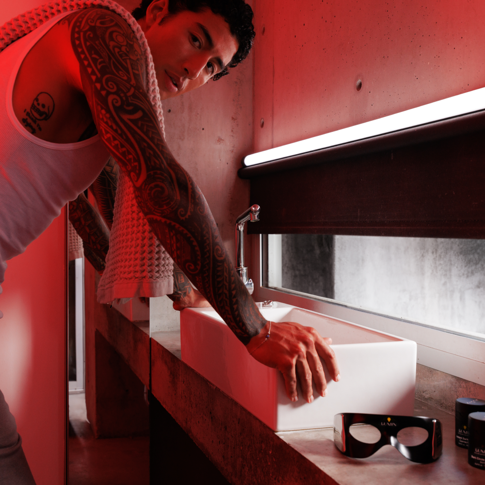 Man with tribal tattoos wearing white tank top and towel washing hands in modern bathroom with concrete surfaces under red lighting