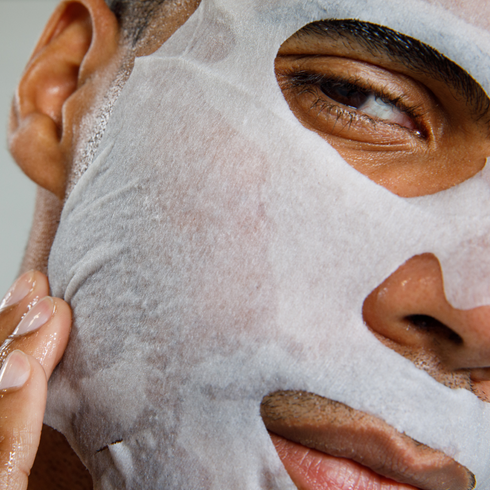 Man applying moisturizing white sheet face mask for skincare routine, promoting hydration and healthy skin care.