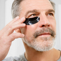 Man applying black under-eye gel patch for skincare and anti-aging treatment, gray beard, home bathroom background.